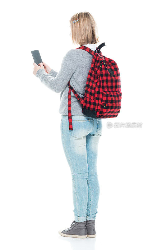 全长/后视图/背面16-17岁美丽的金发z一代年轻女性/少女/学生/高中生/初中生站着学习，拿着手机/包/使用智能手机